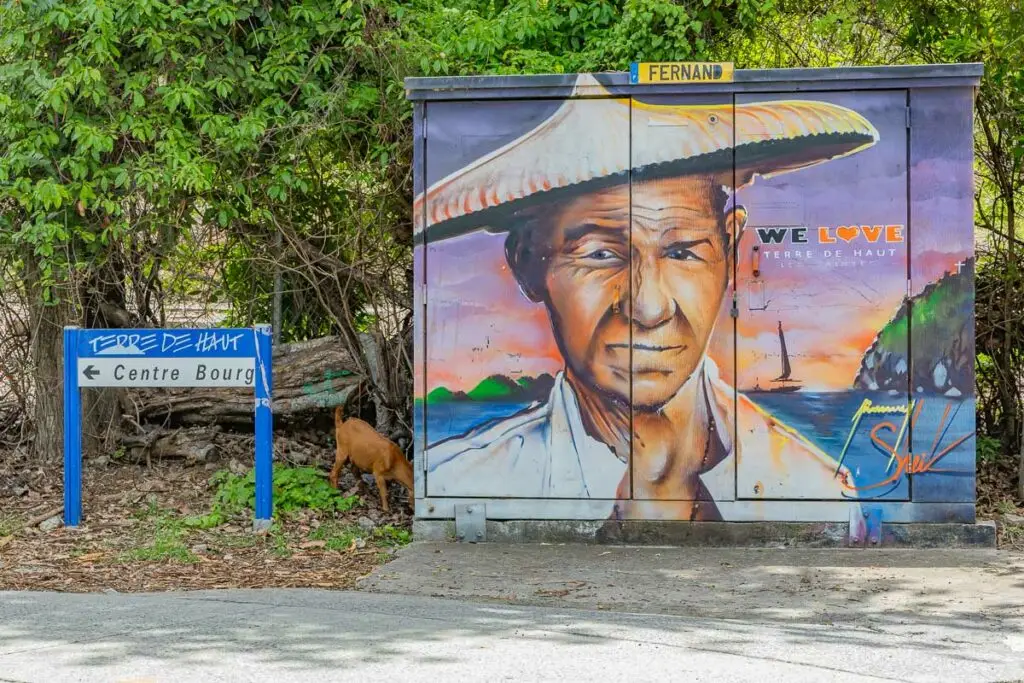 Street art à Terre-de-Haut, Les Saintes, représentant un portrait coloré sur une borne décorée, avec paysage côtier en arrière-plan